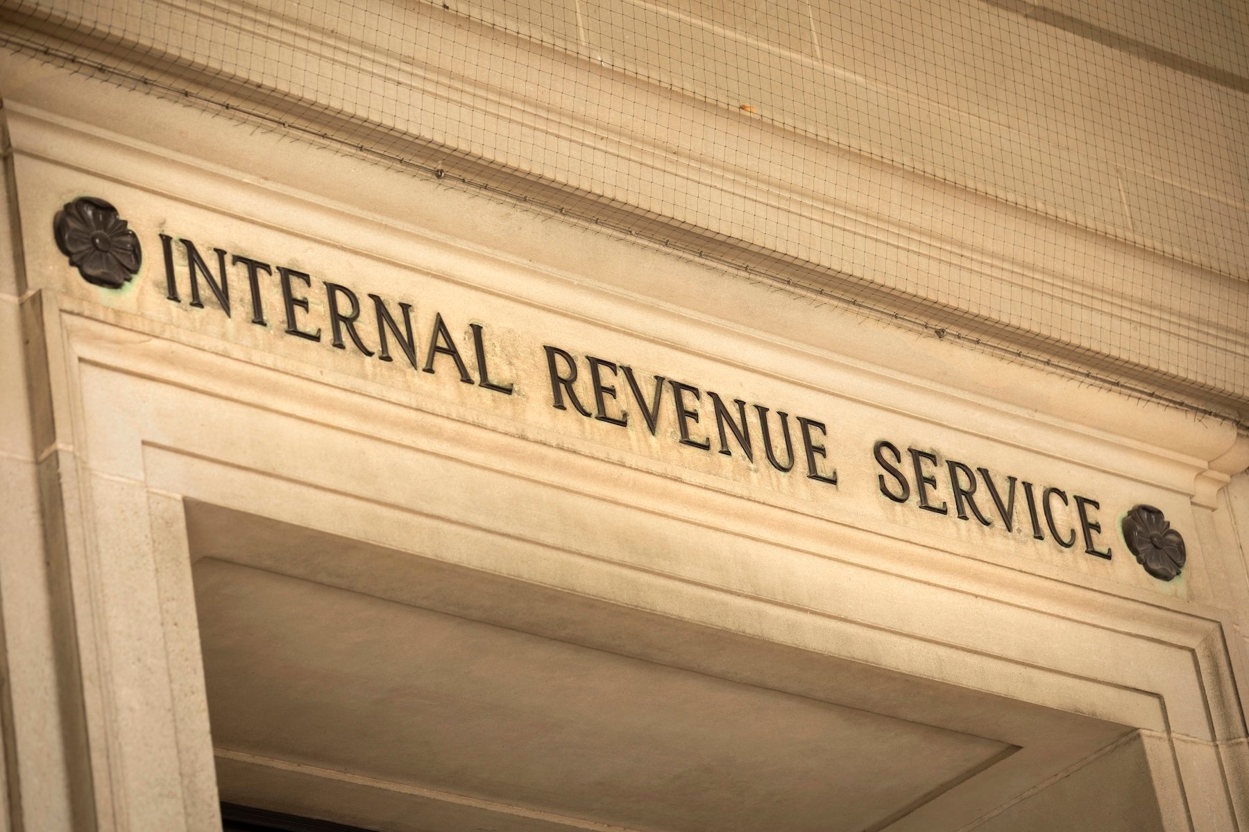 A close-up, slightly upward-angled shot of the Internal Revenue Service (IRS) federal building's entrance in Washington D.C., USA, emphasizing its prominent stone facade and the bold, black carved letters spelling "INTERNAL REVENUE SERVICE" above the doorway. The classic, ornate architectural details, including decorative dark rosettes flanking the text and intricately carved stone trim, are clearly visible. The image conveys a sense of official authority and directly relates to the article discussing the IRS's annual inflation adjustments for retirement accounts, specifically the raised 2026 contribution limits for 401(k) and IRA plans, as detailed in Notice 2025-67.