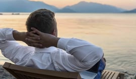 A man with blonde hair, dressed in a white collared shirt, sits relaxed on a wooden bench with his hands clasped behind his head, gazing out over a calm body of water at sunset. A silver laptop is visible next to him on the bench, suggesting he has just finished working or is taking a break while his investments generate passive income. The warm, soft light of the setting sun creates a tranquil atmosphere, emphasizing the freedom and peace of mind associated with achieving financial independence through passive income streams. This image perfectly illustrates the article's core message about earning money without continuous active effort, highlighting the desired outcome of strategic monetary or time investments.