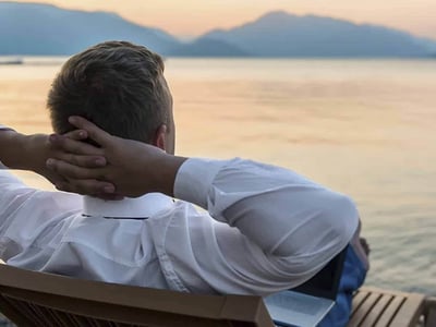 A man with blonde hair, dressed in a white collared shirt, sits relaxed on a wooden bench with his hands clasped behind his head, gazing out over a calm body of water at sunset. A silver laptop is visible next to him on the bench, suggesting he has just finished working or is taking a break while his investments generate passive income. The warm, soft light of the setting sun creates a tranquil atmosphere, emphasizing the freedom and peace of mind associated with achieving financial independence through passive income streams. This image perfectly illustrates the article's core message about earning money without continuous active effort, highlighting the desired outcome of strategic monetary or time investments.