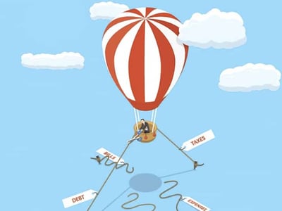 An illustrative image depicting a large, red and white striped hot air balloon ascending into a clear blue sky with fluffy white clouds, representing financial uplift despite burdens. Dangling below the balloon's basket are three separate individuals, each tethered to a significant financial obligation, symbolizing the weight of tax debt and back taxes. The person on the left is holding a sign labeled "BILLS," the central figure is attached to a large gold coin or medallion, and the person on the right is holding a sign clearly marked "TAXES," emphasizing the core theme of the article regarding owing back taxes to the IRS. This visual metaphor effectively conveys the struggle and eventual potential relief associated with managing and overcoming various forms of debt, particularly tax debt. The "The College Investor" logo is visible in the bottom left corner, reinforcing the source of the financial advice.