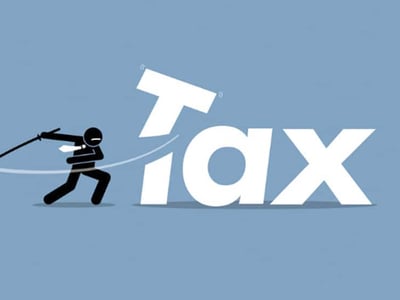 A dynamic cartoon illustration features a stick-figure business person in a black suit and white tie, intensely swinging a katana sword. They are positioned to the left of large, white, bold text that spells out "TAX." The "T" in "TAX" appears to be an uppercase letter that has been cut into two pieces by the sword, with the top portion tilting and falling. The figure's aggressive stance and the fragmented "T" suggest an action of "slaying" or tackling taxes, directly referencing "TaxSlayer" and the concept of overcoming tax preparation challenges. The background is a solid, muted blue, providing a clear contrast for the dark figure and white text. In the bottom left corner, in white sans-serif font, the logo "THE COLLEGE INVESTOR" is visible, indicating the source and context of this image within an article discussing TaxSlayer alternatives and tax software comparisons.