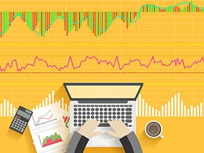 This illustrative image depicts a top-down view of a person using a laptop with hands visible on the keyboard, surrounded by investment-related items on a bright yellow desk. Above the workspace, two distinct stock market charts are displayed against a yellow background, featuring green and red bar graphs and a fluctuating line graph, symbolizing stock performance and trends. To the left of the laptop, a black calculator and a document with a red and green line graph are visible, representing financial calculations and stock analysis. On the right, a cup of coffee suggests a focused work environment. The bottom left corner prominently features the text "THE COLLEGE INVESTOR." This image effectively visualizes the intensive research and analytical work involved in stock picking and investment services like The Motley Fool, as discussed in the accompanying article.