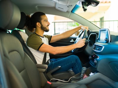 Side view of a smiling young man with tied-back curly hair and a beard, driving a car with a hands-free navigation device mounted on the dashboard. He is dressed in a casual t-shirt and jeans, with his hands comfortably gripping the steering wheel. This image illustrates the flexibility and income potential of driving-based side hustles—such as rideshare, food delivery, and package transport—highlighted in the "10 Proven Ways to Make Money Driving" guide. Source: The College Investor