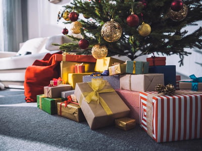 A large pile of wrapped presents under a decorated Christmas tree. This scene illustrates the typical holiday abundance of toys and material gifts, highlighting the alternative value of gifting stock or investments to build a child's future wealth instead of adding to household clutter. Source: The College Investor