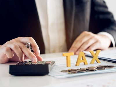 Close-up of a businessman in a suit using a calculator next to coins and wooden letters spelling "TAX." This illustrates the financial planning involved when considering a tax refund advance loan to bridge the gap between filing taxes and receiving a refund from the IRS. Source: The College Investor