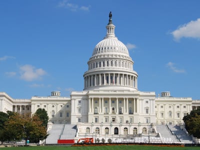 An eye-level, slightly upward-angled, daytime shot of the U.S. Capitol Building in Washington D.C. under a clear blue sky with sparse white clouds. The iconic white dome, topped with the Statue of Freedom, dominates the center of the frame, surrounded by its intricate columns and architectural details. The building's white marble facade extends horizontally, flanked by lush green trees on both sides, with more distant buildings partially visible. In the foreground, scaffolding and construction equipment are visible on the steps leading up to the Capitol, suggesting ongoing work. This image visually represents the "Congress" mentioned in the article, specifically the legislative body responsible for the "Big Beautiful Bill" that eliminates federal Grad PLUS loans, impacting graduate students' ability to afford professional programs like law and medicine.