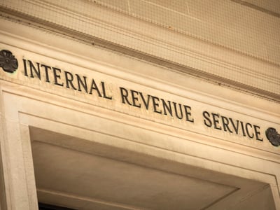 A close-up, slightly upward-angled shot of the Internal Revenue Service (IRS) federal building's entrance in Washington D.C., USA, emphasizing its prominent stone facade and the bold, black carved letters spelling "INTERNAL REVENUE SERVICE" above the doorway. The classic, ornate architectural details, including decorative dark rosettes flanking the text and intricately carved stone trim, are clearly visible. The image conveys a sense of official authority and directly relates to the article discussing the IRS's annual inflation adjustments for retirement accounts, specifically the raised 2026 contribution limits for 401(k) and IRA plans, as detailed in Notice 2025-67.