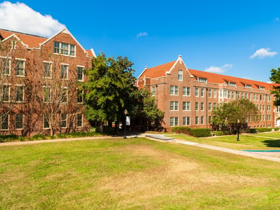 A bright, sunny view captures two imposing traditional red-brick college dormitory buildings with distinctive clay-tiled roofs, set against a vibrant blue sky with scattered white clouds. The multi-story dormitories feature numerous windows and are nestled within a well-maintained campus landscape, complete with manicured green lawns that sweep across the foreground and extend between the buildings. Several mature deciduous trees with lush green foliage are interspersed throughout the scene, casting soft shadows. A paved pathway winds through the grass, leading towards the dorm entrances. This visual representation highlights typical on-campus housing facilities, emphasizing the "room and board" expense which is a significant, and often overlooked, financial component of college costs that can even exceed tuition fees at many public universities. Source: The College Investor