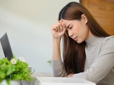 A side view of a young Asian woman with long brown hair, visibly stressed and overwhelmed, sitting at a light wooden desk. Her eyes are closed, her brow furrowed, and her right hand is pressed against her forehead, holding a dark pencil, indicating deep frustration or fatigue. A black laptop is open on the left side of the desk, along with a white coffee mug and a small green plant with white flowers, suggesting a home or office study environment. The background is a blurred light wall and a wooden door. This image powerfully illustrates the immense academic pressure, financial hardship, and mental health struggles that contribute to the alarming college dropout rate, where nearly 40% of students leave without earning a degree due to affordability issues, academic difficulties, and personal responsibilities. Source: The College Investor