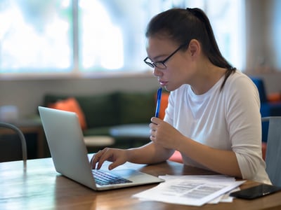 A focused young woman, wearing glasses and a white t-shirt, sits at a wooden table intently working on a silver laptop, representing the diligent process of researching and managing private student loans. She holds a blue pen to her chin in a thoughtful pose, with several papers and a dark smartphone resting beside her, indicating the detailed financial documents and information involved in student loan decisions. The background, blurred to emphasize her concentration, shows a modern indoor setting with large windows and hints of green and orange furniture, suggesting she could be in a college library, co-working space, or home office. This image visually conveys the careful consideration and analysis required when families, particularly parent borrowers, weigh federal versus private student loan options amidst changing repayment programs and interest rates for college funding. | Source: The College Investor