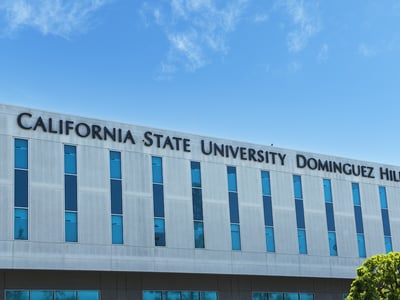 A wide, eye-level shot on a bright, clear day shows the modern, light-gray building of California State University Dominguez Hills under a blue sky with scattered wispy clouds. The university's name, "CALIFORNIA STATE UNIVERSITY DOMINGUEZ HILLS," is prominently displayed in large, dark blue, sans-serif letters across the top facade of the building. Below the signage, multiple rows of tall, rectangular windows with dark frames and reflective blue glass are evenly spaced, creating a pattern across the building's surface. In the bottom right corner, the vibrant green foliage of a tree is partially visible, adding a touch of nature to the academic setting. This image visually represents one of the California State University (CSU) campuses, providing context for the article's discussion of the new California law, Senate Bill 640 (SB 640), which will implement the Direct Admission Program for qualified California high school seniors starting in 2027. The law aims to boost college enrollment and ease the application process for students.