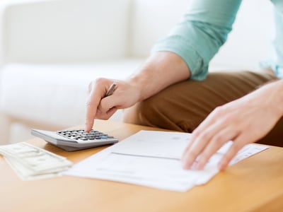 A person in a light blue shirt and khaki pants sits at a light wooden table, actively calculating figures with a silver and black calculator. Their right hand points a finger at the calculator keys, while their left hand rests on a stack of white papers, possibly bills or financial documents. To the left of the calculator, a small stack of US dollar bills, likely twenty-dollar denominations, is visible, suggesting money management or financial planning. The background is softly blurred but shows a white sofa, indicating a home or office setting. This image visually represents the process of managing finances and calculating costs, directly relating to the article's focus on understanding and comparing student loan rates to save borrowers money over time. Source: The College Investor
