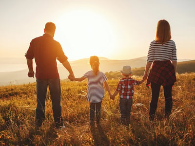 A family of four, including two adults and two young children, stands hand-in-hand facing away from the viewer, looking out over a sun-drenched, golden field at sunset or sunrise. The father, on the left, wears a plaid shirt and jeans, holding his daughter's hand. The daughter, in a striped shirt, holds her younger brother's hand, who is wearing a plaid shirt and a straw hat. The mother, on the right, wears a striped shirt, dark pants, and a plaid shirt tied around her waist, holding her son's hand. The warm, golden light of the sun, positioned centrally in the background, casts long shadows and illuminates the family and the expansive landscape of rolling hills and mountains in the distance. This image visually represents the "Family of Four Middle Class Squeeze" discussed in the article, highlighting the challenges middle-class families face with rising living costs and benefit cliffs, despite earning $100,000 to $200,000, as they struggle to get ahead financially. Source: The College Investor