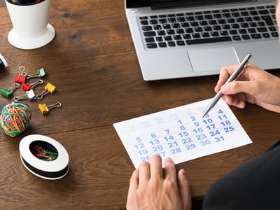 An overhead shot captures a person's hands holding a silver pen over a printed calendar page, likely marking or planning for the upcoming July 1, 2026 deadline. The calendar, displaying the days of a month, rests on a warm, dark wooden desk. To the right, a silver laptop with a visible keyboard and touchpad is open, suggesting a work or research environment. Various office supplies are scattered around the desk, including a white pen holder with a dark pen, a small pile of colorful binder clips, a multi-colored rubber band ball, and a white oval dish filled with paper clips. This detailed scene visually represents the meticulous planning and organization required as individuals and families prepare for the significant federal policy changes impacting student loan caps and repayment options beginning in 2026. Source: The College Investor