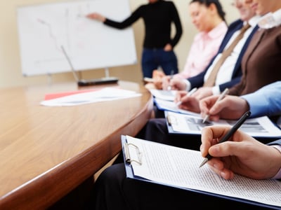 A blurry, close-up shot of a group of professionals attending a training or briefing session in an office conference room, underscoring the article's focus on Training Repayment Agreement Provisions (TRAPs). In the foreground, a person's hand is visible, holding a black pen and writing notes on a document attached to a clipboard, emphasizing the active learning and contractual nature of job training. Other attendees are partially visible in the middle ground, also taking notes, with one individual in a light pink shirt and another in a brown top. In the background, out of focus, a presenter in a black shirt gestures towards a whiteboard, which has diagrams or notes written on it, symbolizing the "training costs" discussed in the article. The warm wooden conference table dominates the left side of the frame, grounding the professional setting. This image visually represents the formal training environments where TRAPs are often implemented, highlighting the contracts and agreements workers sign that can lead to significant debt if they leave their employment early. | Source: The College Investor