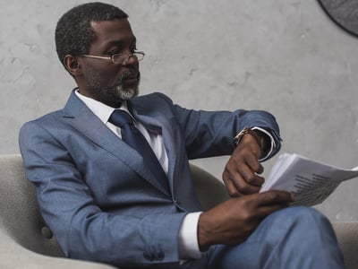 A distinguished Black man in a tailored blue suit and white shirt, accented with a dark patterned tie, sits in a modern gray armchair against a textured concrete wall. He wears sophisticated gold-rimmed glasses and has neatly groomed gray hair and a beard. His expression is one of focused anticipation as he holds a stack of papers, seemingly reports or applications, in his hands, looking down. This image effectively illustrates the article's theme of "Waiting on a report" regarding the Public Service Loan Forgiveness (PSLF) Buyback backlog, reflecting the ongoing delays and the pause in monthly status reports due to the government shutdown, leaving over 74,000 pending applications in limbo for borrowers awaiting updates on their student loans.