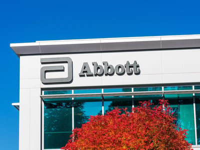 This image features the prominent gray logo and wordmark "Abbott" on the clean, white facade of an Abbott Laboratories building, signifying the company's corporate identity. Below the logo, a striking tree with vibrant red and orange autumn foliage adds a natural contrast against the modern architecture and the clear, bright blue sky above. The building's windows reflect the sky and surrounding environment, adding depth to the scene. This visual serves to contextualize Abbott Laboratories as one of the major U.S. employers offering retirement plan matches tied to student loan payments, a key point discussed in the accompanying article about the SECURE 2.0 provision. Source: The College Investor