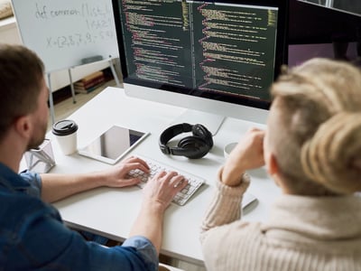 A high-angle, over-the-shoulder shot shows two young professionals, likely Gen Z, intensely focused on a computer monitor displaying lines of code. The male on the left, wearing a denim shirt, types on a white keyboard, while the female on the right, with blonde hair pulled back in a bun and wearing a cream-colored ribbed sweater, observes. The brightly lit white desk features a black coffee cup, a white tablet, and black over-ear headphones, suggesting a modern workplace or collaborative environment. In the background, a whiteboard with handwritten notes adds to the coding context. This image visually represents the contemporary, tech-driven work settings where Gen Z individuals are making financial decisions like withdrawing from 401(k) or IRA accounts due to rising living costs, as discussed in the accompanying article on Gen Z's financial behaviors. Source: The College Investor