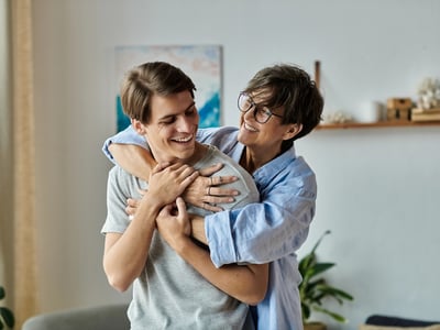 A loving mother with short, dark, salt-and-pepper hair and stylish round glasses, wearing a light blue button-down shirt, embraces her adult son from behind in a warm, brightly lit home. Both are smiling broadly, looking at each other affectionately, reflecting a close family relationship. The son, with short brown hair and a grey t-shirt, is facing forward, his hands resting gently on his mother's arms wrapped around him. This image illustrates the close bond between parents and adult children, which is central to the article discussing why shared bank accounts can create legal, tax, and financial aid risks as children become adults. The cozy home setting features blurred background elements like a light-colored wall, a painting with blue hues, and a plant, suggesting a comfortable, personal environment where financial discussions like those in the article might take place. Source: The College Investor