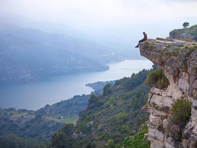 A lone figure sits on the jagged edge of a high cliff overlooking a misty river valley. This visualizes the "student loan tax cliff" approaching on December 31, 2025, when the federal tax exemption for loan forgiveness expires. The precarious setting symbolizes the urgency for borrowers, particularly those on the SAVE plan, to act quickly to secure their forgiveness status before facing a potential tax bill. Source: The College Investor