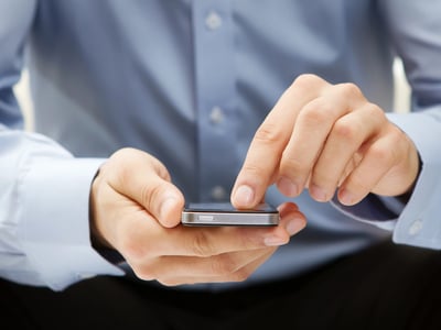 A close-up shot shows a person wearing a light blue dress shirt, with the sleeves rolled up, holding a silver smartphone horizontally with both hands. Their thumbs are positioned over the screen, poised to interact with the device, which glows faintly. This image directly relates to the article's discussion of the "vice economy," where mobile gambling and sports betting apps blur the lines between entertainment and finance for young adults. The phone represents the ease of access to these activities, contrasting with traditional financial advice and highlighting how social media, betting apps, and speculative investing contribute to a culture of instant gratification. Source: The College Investor