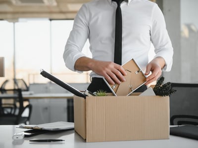 A close-up shot of a businessperson, dressed in a white shirt and black tie, carefully placing personal items into a cardboard box on a white office desk. The person's face is not visible, emphasizing the act of packing. Inside the box, a picture frame, a small potted plant, and decorative black berries are discernible, suggesting a recent termination or "tech layoff" as discussed in the article. The background shows a modern office environment with large windows, blurred office chairs, and desks, indicating a corporate setting. This image powerfully illustrates the current struggles faced by young workers in the tech industry, including college graduates and soon-to-be graduates, who are experiencing fewer job opportunities and a tougher path into the sector due to mass layoffs and a reshaping of early-career hiring patterns. Source: The College Investor