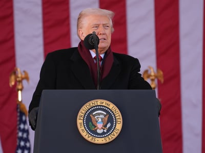 President Donald Trump, with his signature blonde hair, stands authoritatively at a dark podium adorned with the official Seal of the President of the United States. He is dressed in a smart black overcoat and a rich maroon scarf, speaking into a microphone positioned on the podium. Behind him, large American flags with prominent red and white stripes create a patriotic backdrop, flanked by golden eagle finials atop flagpoles. This image visually represents the executive branch's influence, illustrating the context of the article discussing the Trump administration's efforts to block the Consumer Financial Protection Bureau (CFPB) from accessing Federal Reserve funds, a move that could deplete the agency's budget by early 2026 and potentially lead to its effective closure. (AP Photo/Evan Vucci)