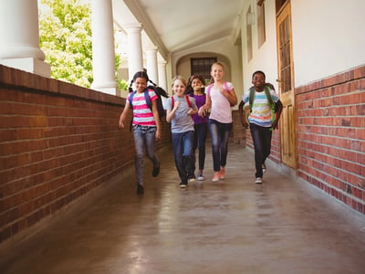 Smiling elementary school students with backpacks running down an outdoor school corridor lined with brick walls and white columns. This image illustrates the expanded utility of 529 plans, which can now be used for a broader range of K-12 educational expenses, including tuition, tutoring, and materials, with annual caps rising to $20,000 in 2026. Source: The College Investor