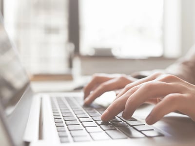 Close-up of a student’s hands typing on a silver laptop keyboard next to textbooks and glasses. This image visualizes the urgency and focus required when applying for mid-year student loans or an Education Line of Credit to cover unexpected college expenses like housing or course fees after the semester has already begun. Source: The College Investor