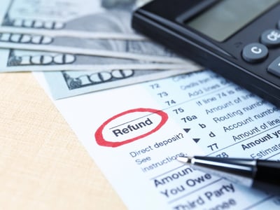 Close-up of a calculator, U.S. hundred-dollar bills, and a tax form with the word "Refund" circled in bold red ink. This image highlights the projected increase in 2026 tax refunds due to retroactive tax law changes, such as new deductions for tip income, overtime pay, and auto loan interest, which were not reflected in paycheck withholding throughout the year. Source: The College Investor