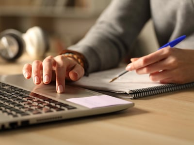 Close-up of a student’s hands typing on a laptop and taking notes in a spiral notebook, with headphones in the background. This image illustrates the college essay writing process, highlighting the tension students face between using digital tools like AI for assistance and ensuring their work reflects their own authentic voice, a key concern for admissions officers. Source: The College Investor