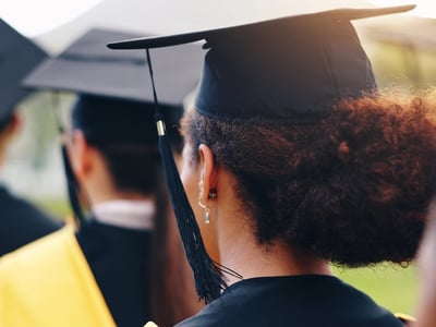Rear view of a row of diverse college graduates wearing black caps and gowns with gold tassels during an outdoor commencement ceremony. The image represents the 61.1% of students from the fall 2019 cohort who successfully completed their degree within six years, illustrating the steady but flat national graduation rate highlighted in the latest data. Source: The College Investor