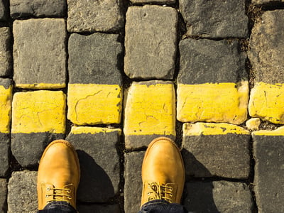 Person's feet in yellow boots standing at a bright yellow line painted across gray cobblestones. This visual metaphor represents the critical threshold facing graduate students: the hard cutoff date of June 30, 2026, marking the end of the Grad PLUS loan program and the start of strict new federal borrowing caps. Source: The College Investor