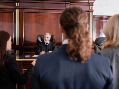 A judge in black robes listens intently to two attorneys arguing their case in a wood-paneled courtroom. This legal setting illustrates the professional sectors—specifically law, medicine, and dentistry—most impacted by the new 2026 federal student loan caps, which limit annual borrowing to $50,000 for professional degrees. Source: The College Investor