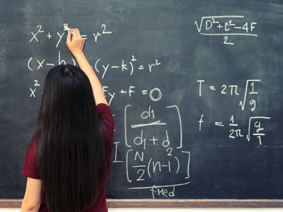 Rear view of a female student with long dark hair, wearing a maroon shirt, writing complex algebraic equations and geometry formulas on a large chalkboard. This image illustrates the critical role of mastering high school math concepts—specifically Algebra 2 and Geometry—as foundational preparation for success on the SAT and ACT exams. Source: The College Investor