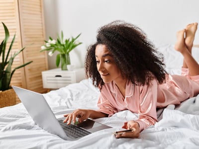Young woman with natural curls lying on her stomach in bed, wearing satin pink pajamas and browsing on a laptop while holding a credit card. This image illustrates the seamless integration of digital finance into daily life, reflecting 2025 trends where consumers increasingly manage payments, banking, and investments online from the comfort of home. Source: The College Investor