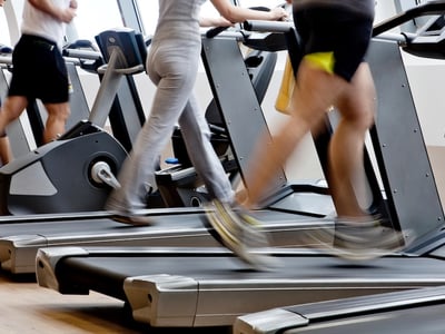 Motion-blurred legs of people walking and running on treadmills in a bright gym. This image illustrates the common New Year's resolution of improving physical fitness, while serving as a visual hook for discussing the financial costs of gym memberships and offering budget-friendly alternatives for 2026 health goals. Source: The College Investor