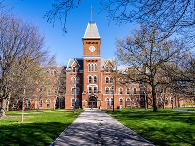 A red brick University Hall at Ohio State University featuring a distinctive clock tower stands at the end of a tree-lined walkway under a clear blue sky. This classic collegiate setting illustrates the traditional "sticker price" perception of college, while the accompanying article explains how students can avoid paying full tuition through grants, community college transfers, and employer-paid programs.