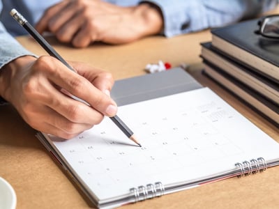 Overhead shot of a person’s hands holding a pencil, pointing to a specific date on a paper calendar laid flat on a wooden desk. Beside the calendar are a coffee cup, laptop, and a stack of books with glasses on top. This image illustrates the crucial planning needed for Parent PLUS loan borrowers, who face a strict consolidation disbursement deadline of June 30, 2026, to remain eligible for income-driven repayment plans like ICR and IBR. Source: The College Investor