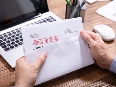 Overhead shot of a person’s hands opening a white envelope stamped with the words "FINAL NOTICE" in bold red letters. Beside the document sits a laptop and other office items on a wooden desk. This image visually represents the financial stress and looming deadlines facing student loan borrowers, highlighting the surge in delinquencies and the potential restart of payments for millions following recent policy shifts. Source: The College Investor