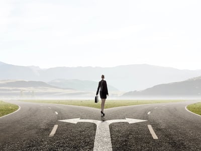 Businesswoman standing at a fork in a rural road, contemplating two distinct paths that stretch toward distant mountains. This visual metaphor represents the strategic choice families face when deciding between the specialized education savings of a 529 Plan and the broader, long-term financial flexibility of the newly proposed Trump Accounts. Source: The College Investor