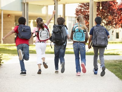 Diverse group of smiling elementary school children running away from the camera down a sunny walkway lined with trees and school buildings. The children are wearing backpacks, representing the broad range of K-12 students who could benefit from the new Education Freedom Tax Credit, which aims to fund scholarships for public, private, and charter school expenses. Source: The College Investor