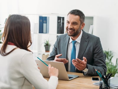 A smiling businessman in a grey suit and blue tie gestures while speaking to a client in a bright office. This image represents the role of financial professionals—specifically Certified Financial Planners (CFPs) or student loan consultants—who provide strategic advice to borrowers navigating complex repayment options, forgiveness plans, and long-term financial goals. Source: The College Investor
