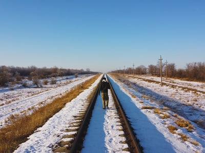 Lone figure walking away down the center of a snow-covered railroad track stretching toward the horizon under a clear blue sky. This visual metaphor represents the narrowing path and strict deadlines facing Parent PLUS loan borrowers in 2026, as they navigate the critical window for consolidation and repayment options before the tracks effectively "end" for new loans. Source: The College Investor