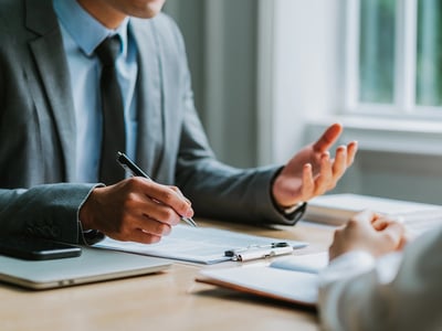 Close-up of a businessman in a gray suit holding a pen while explaining a contract to a client at a wooden table. This image illustrates the consultation process for refinancing private student loans, specifically highlighting the new option of income-based repayment plans offered by lenders like RISLA. Source: The College Investor