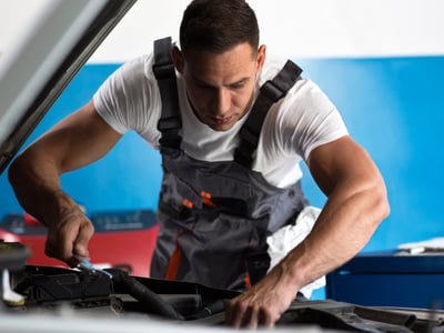 Close-up of a focused mechanic wearing a white T-shirt and grey overalls working under the open hood of a car, holding a wrench. This image serves as a visual metaphor for the student loan rehabilitation process—a deliberate, hands-on repair strategy that allows borrowers to fix their defaulted loans and restore their financial standing through consistent effort. Source: The College Investor