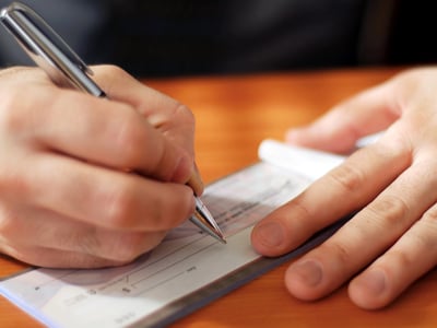 Close-up of a person's hands writing a check on a wooden desk. This image illustrates the reality of out-of-pocket college costs, where most families end up paying between $25,000 and $100,000 for a bachelor's degree after financial aid is applied. Source: The College Investor
