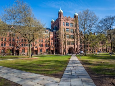 Sunny view of a paved pathway leading toward the historic, castle-like red brick buildings of Yale University's campus. This image visually anchors the news of Yale raising its free-tuition income threshold to $200,000, highlighting the elite institution's efforts to expand affordability amidst rising costs. Source: The College Investor