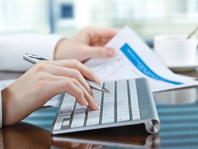 Close-up of a person’s hands typing on a modern keyboard next to financial documents and a cup of coffee. This image illustrates the process of managing tax information, specifically focusing on Form 1098-T, scholarships, and understanding the tax implications for students and families. Source: The College Investor