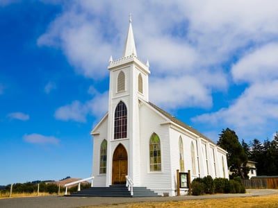 Traditional white wooden church with a tall steeple and arched stained-glass windows, standing against a bright blue sky with white clouds. This image represents the analysis of educational attainment among U.S. religious groups, specifically highlighting the diverse range of college completion rates within Christian denominations, from Episcopalians to evangelicals. Source: The College Investor